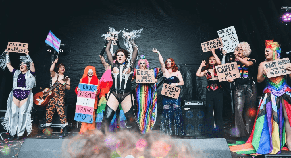 A group of performers in bright clothes holding protest signs during a gay pride event. 