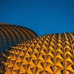 A photograph of a lattice work building structure against a blue sky background. The lattice work is formed from complex interlocking geometric shapes.